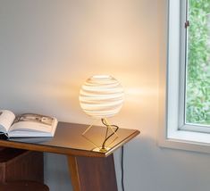 a table with a lamp on it next to an open book and a window in the background