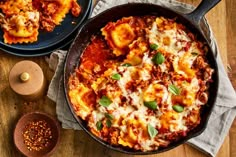 two skillets filled with pasta and sauce on top of a wooden table next to a bowl of seasoning