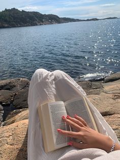 a woman is reading a book on the rocks by the water with her hand resting on an open book