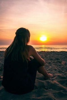 a woman sitting on top of a sandy beach next to the ocean in front of a sunset