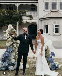 a bride and groom holding hands in front of a large white house with blue flowers