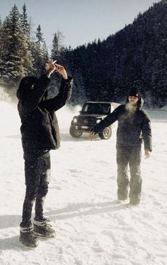 two men standing in the snow next to a car and one is holding something up