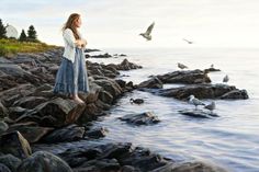 a painting of a woman standing on rocks near the ocean with seagulls flying overhead