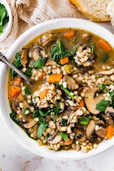 a white bowl filled with mushroom barley soup next to a slice of baguette bread