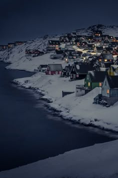 a small town is lit up at night by the water's edge with snow on the ground