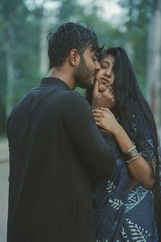 a man and woman standing next to each other in front of some trees with their faces close together