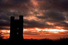 the sun is setting behind a tower with clouds in the sky and trees silhouetted against it