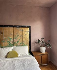 a bed with white sheets and pillows next to a potted plant on a nightstand