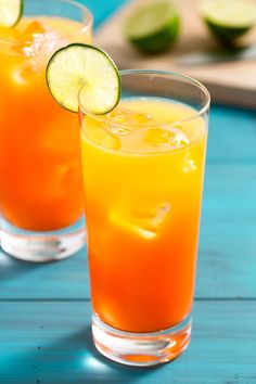 two glasses filled with orange juice and limes on top of a blue table next to a cutting board