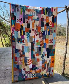 a multicolored quilt hanging on a clothes line in front of a tree and dirt road