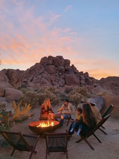 four people sitting around a fire pit in the desert at sunset with rocks behind them