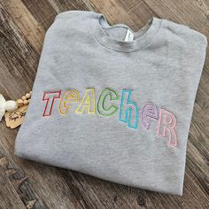 Welcome to my shop! This listing is for an embroidered crewneck sweatshirt!  *Sweatshirt is a Bella Canvas drop shoulder pullover in heather gray. Sizing runs unisex, these are a high quality sweatshirt with long sleeves and torso (see size chart in photos). Other sweatshirt colors may be done upon request for an additional fee. *Front design is TEACHER in rainbow colors!  Feel free to message me for any custom requests! Can change any wording or colors! PaigesStitches Shop Notes: Thank you for Gray Crew Neck Sweatshirt With Embroidered Graphics, Customizable Crew Neck Sweatshirt For School, Gray Letter Embroidery Sweatshirt For Fall, Crew Neck Sweatshirt With Embroidered Graphics For School, Graphic Embroidered Crew Neck Sweatshirt For School, Embroidered Crew Neck Sweatshirt For School, School Sweatshirt With Embroidered Text And Crew Neck, School Crew Neck Sweatshirt With Embroidered Text, Embroidered Gray Crew Sweatshirt