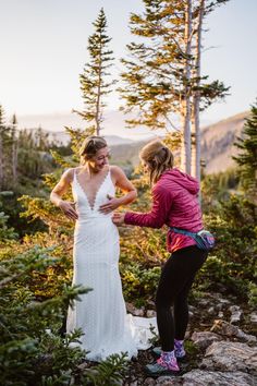 two women standing next to each other on top of a rocky hillside with pine trees in the background