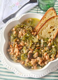 a white bowl filled with beans and greens next to a slice of bread on top of a table