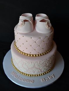 a pink cake with baby shoes on top and the words welcome baby written in gold