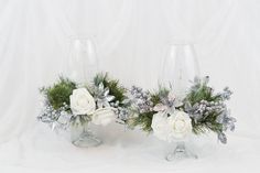 two glass vases with flowers and greenery in them sitting on a white surface