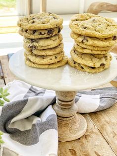 chocolate chip cookies stacked on top of each other on a white cake plate next to succulents