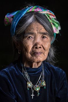 an old woman wearing a blue shirt and a colorful head scarf is looking at the camera