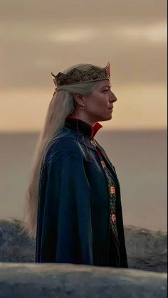 a woman with long blonde hair wearing a crown and standing in front of the ocean