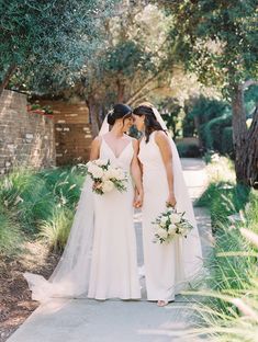 two women in white dresses standing next to each other on a sidewalk with trees and bushes behind them