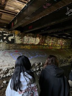 two people standing in front of graffiti on the side of a wall under a bridge