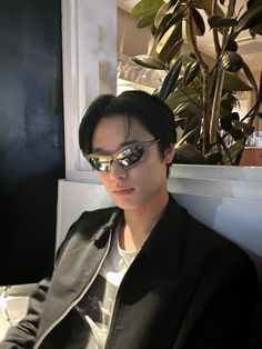 a young man wearing mirrored sunglasses sitting in front of a potted plant and looking off to the side