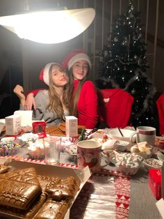 two women sitting at a table covered in food and christmas decorations, with a lit up tree behind them