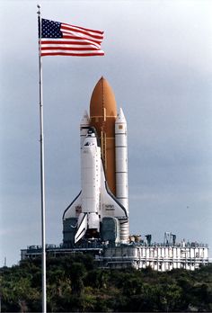 an american flag flies next to the space shuttle on top of a hill with a building in the background