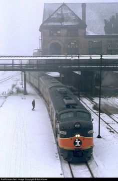 a train traveling down tracks next to a snow covered field with buildings in the background