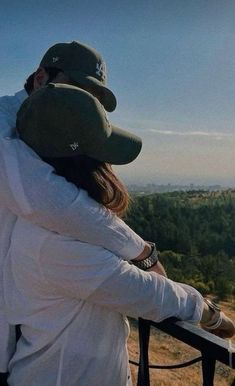 two people standing on top of a hill with trees in the background and one person wearing a hat