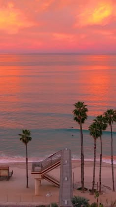 the sun is setting over the ocean with palm trees and benches in front of it