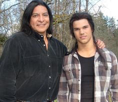 two young men standing next to each other in front of trees and grass, one is wearing a plaid shirt