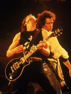 two men playing guitars on stage with one holding the other's neck and looking at something