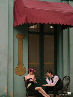 a man and woman sitting at a table in front of a building with a red awning