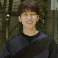 a young man wearing glasses standing in front of a glass wall with plants behind him