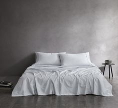 a bed with white sheets and pillows against a gray wall next to a small table