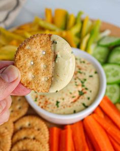 a cracker being dipped with dip surrounded by carrots and cucumbers