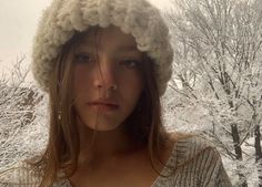 a woman wearing a knitted hat in front of trees with snow on the ground