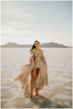 a woman standing in the sand with her dress blowing in the wind and mountains in the background