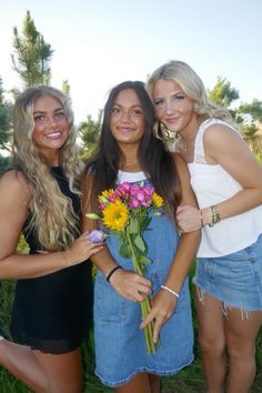 three beautiful young women standing next to each other in front of trees and grass, one holding a bouquet of flowers