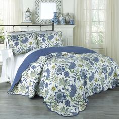 a bed with blue and white flowers on it in front of a mirror, dresser and window