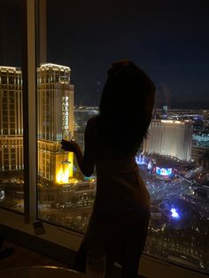 a woman standing in front of a window looking out at the city lights and buildings