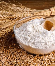 a wooden spoon in a glass bowl filled with flour next to ears of wheat on a table