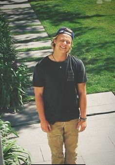 a young man standing on the sidewalk in front of some trees and grass with his hands in his pockets