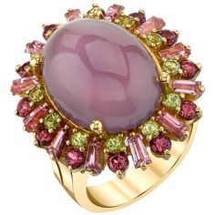 A large, 13.56 carat lavender chalcedony cabochon is featured in this one-of-a-kind cocktail ring. Fine quality, high domed and richly colored lavender chalcedony is rare, especially in large sizes. Pink tourmaline tapered baguettes, mulberry rhodolite garnets and apple green peridots encircle the chalcedony. This selection of pastel gemstones create a colorful frame for the chalcedony reminiscent of an Impressionistic masterpiece. Made of 18k yellow gold by our artisans in Los Angeles. This fun Lavender Cocktail, Month Gemstones, Yellow Gold Cocktail Ring, Gold Cocktail Ring, Gold Cocktail, Gemstone Engagement, Green Gems, Rhodolite Garnet, Green Peridot