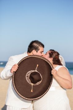 a bride and groom kiss on the beach while holding a large brown hat in front of them