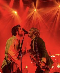 two men singing into microphones on stage with red lights in the backround