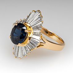 This lovely ring is centered with one (1) oval mixed cut natural sapphire set into a four-prong setting. The sapphire is bordered with twenty-eight (28), channel/prong set, tapered baguette cut diamonds. The ring measures 22.9mm at the top, rises 11.0mm above the finger, tapering to 2.7mm wide and 1.0mm thick at the base of the shank. It is currently a size 6.25. Timeless Oval Sapphire Ring, Timeless Oval Sapphire Ring With Prong Setting, Art Deco Oval Sapphire Ring With Prong Setting, Oval Diamond Cut Sapphire Ring Fine Jewelry, Formal Oval Sapphire Ring With Prong Setting, Elegant Sapphire Oval Cluster Ring, Oval Sapphire Ring With 17 Jewels For Anniversary, Art Deco Oval Sapphire Ring For Anniversary, Classic Oval Sapphire Cluster Ring