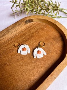 two white earrings with orange and black designs on them sitting on a wooden tray next to a plant