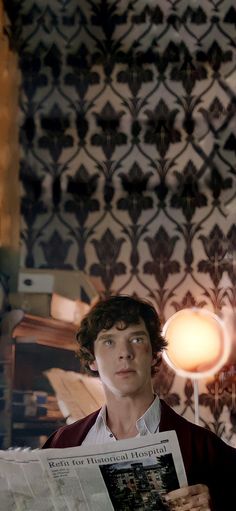 a young man reading a newspaper in front of a wallpapered room with a lamp